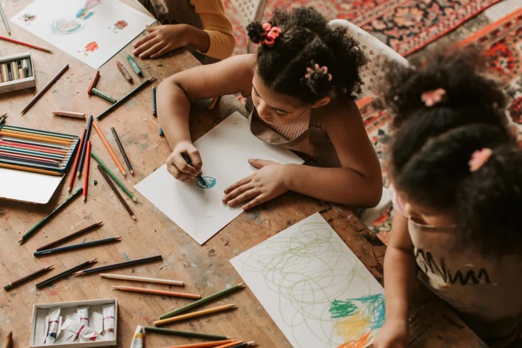 Two girls are pictured from above using crayons and pencils and paper on a table to make drawings.
