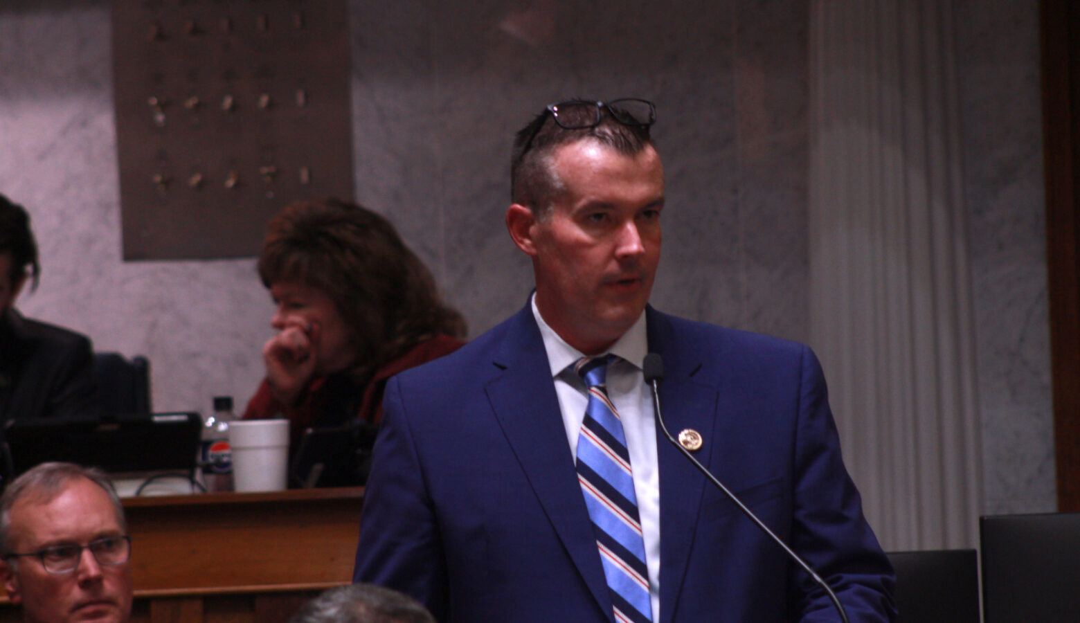 Aaron Freeman stands at the Statehouse, speaking into a long, narrow microphone. He wears a blue suit and his glasses sit on the top of his head.