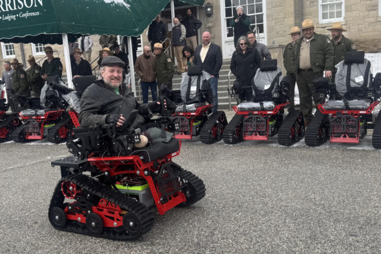A man tries the new red electric track wheelchair in a parking lot, where many people watch him operate it.