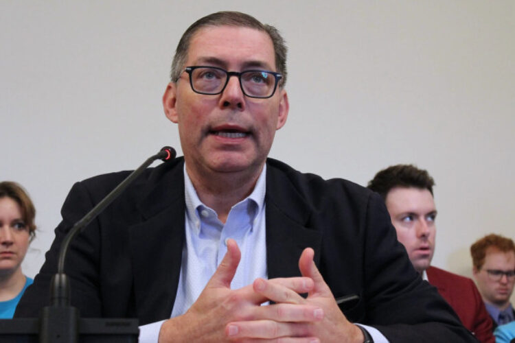 Mitch Roob, Secretary of the FSSA, has requested changes to a federal drug program that health centers warn will cost them millions. He is seen sitting at a table with a microphone speaking to a committee. Spectators are seated behind him in this file photo.