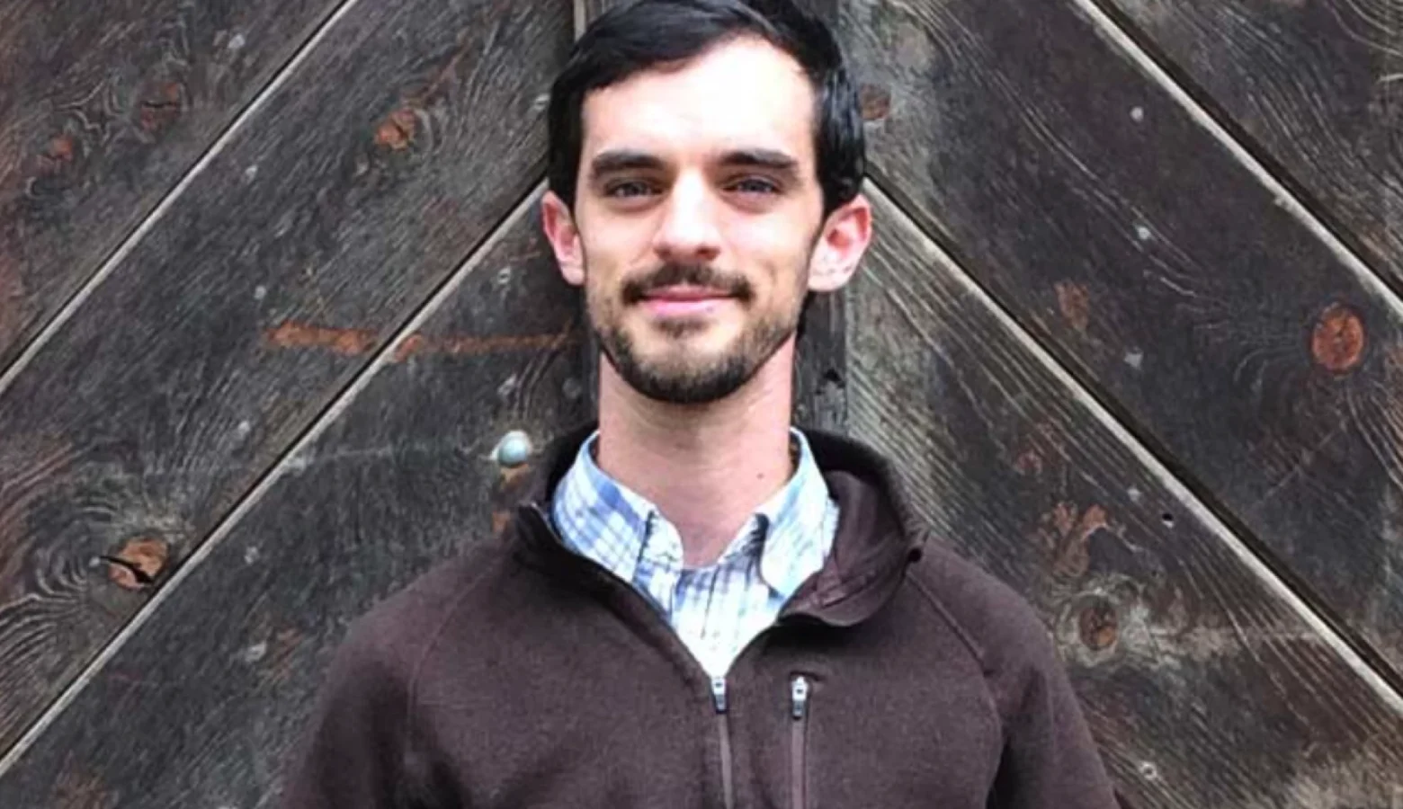 A white man with dark hair and a beard wears a blue checked shirt and a brown fleece jacket.