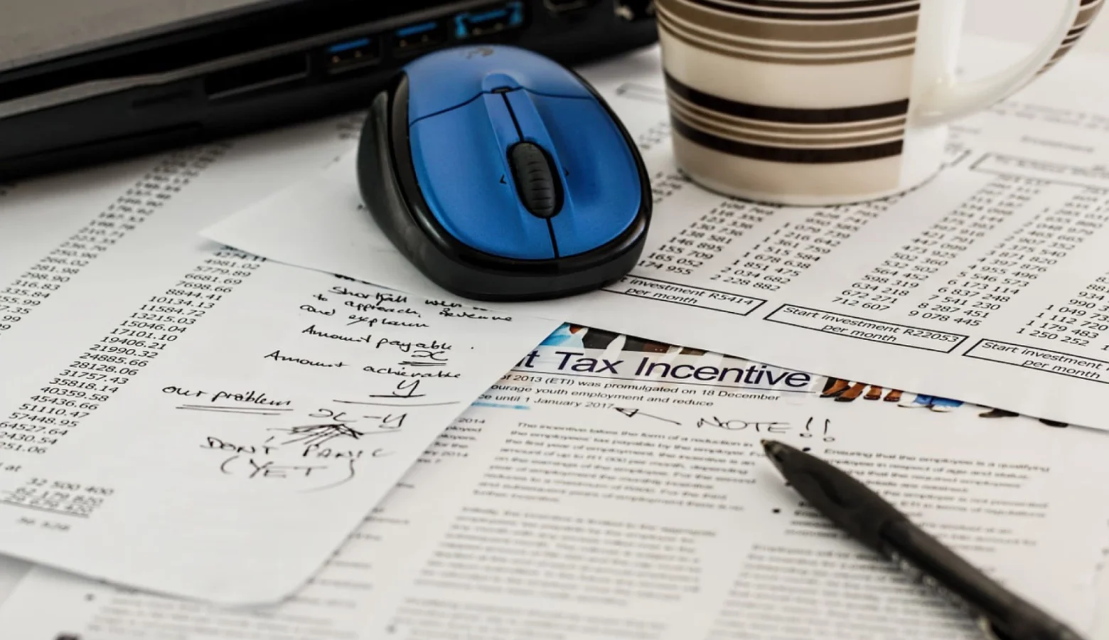 Tax paperwork sits haphazardly across a desk. On top is a blue computer mouse, a black pen, and a coffee mug.