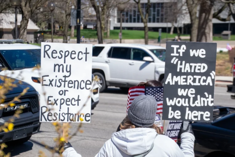 A protester standing on a street is seen from behind, holding two signs. One reads Respect my existence or expect resistance. The other sign reads If we hated America, we wouldn't be here.