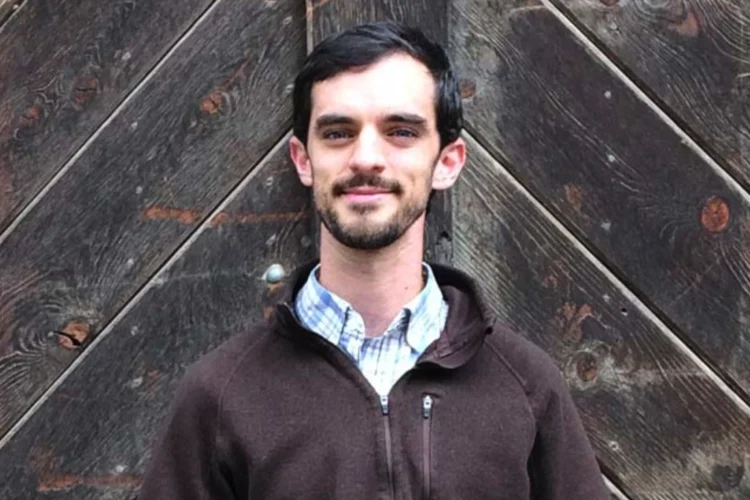 A white man with dark hair and a beard wears a blue checked shirt and a brown fleece jacket.