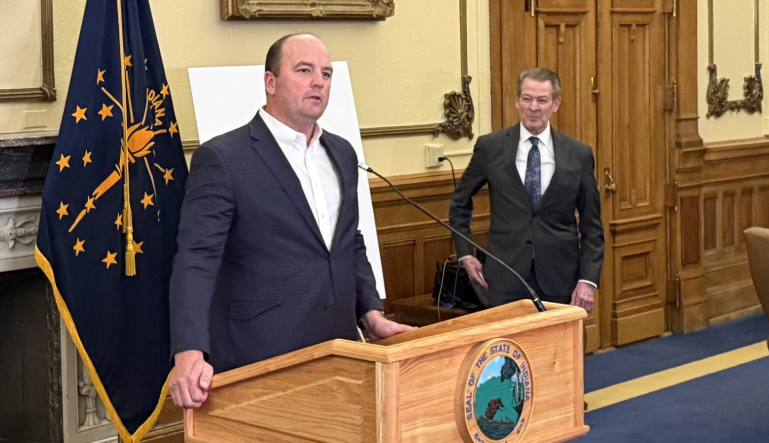 Fishers Mayor Scott Fadness stands at a podium in front of a microphone. He is a bald white man, wearing a white shirt and navy suit jacket.