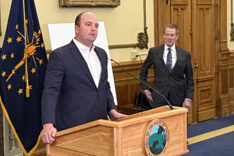 Fishers Mayor Scott Fadness stands at a podium in front of a microphone. He is a bald white man, wearing a white shirt and navy suit jacket.