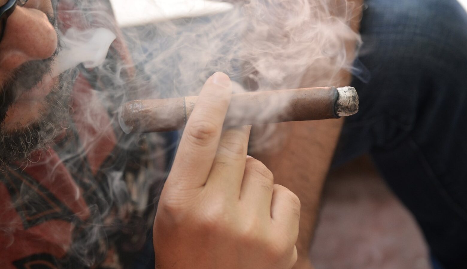 A nearly unseen person holds up a cigar as smoke wafts through the frame.