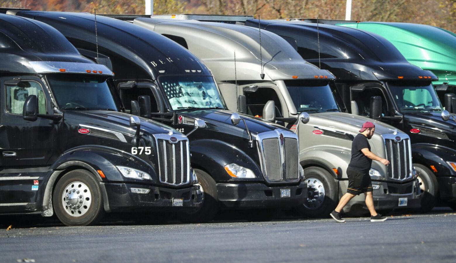 Five semi-trucks are parked and lined up at a truck stop in New Jersey. A driver is walking away from the trucks.