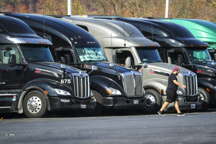 Five semi-trucks are parked and lined up at a truck stop in New Jersey. A driver is walking away from the trucks.