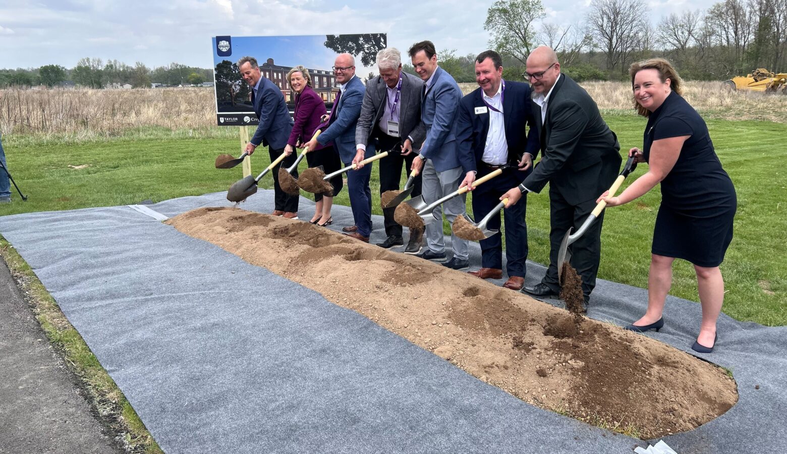 Several people in business attire stand behind a small mound of dirt. They all hold shovels having just scooped up some dirt.