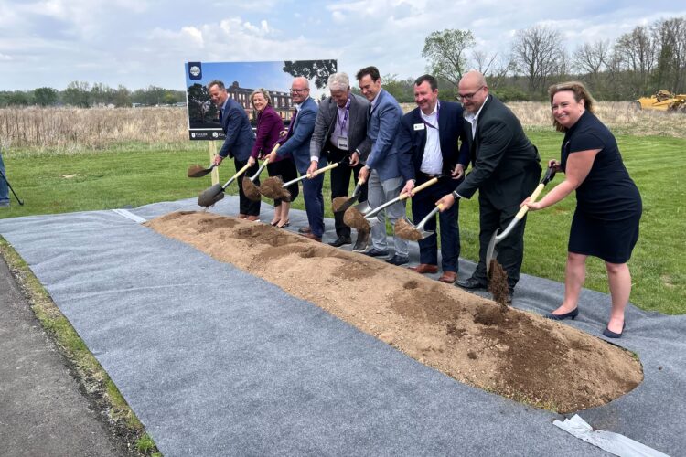 Several people in business attire stand behind a small mound of dirt. They all hold shovels having just scooped up some dirt.