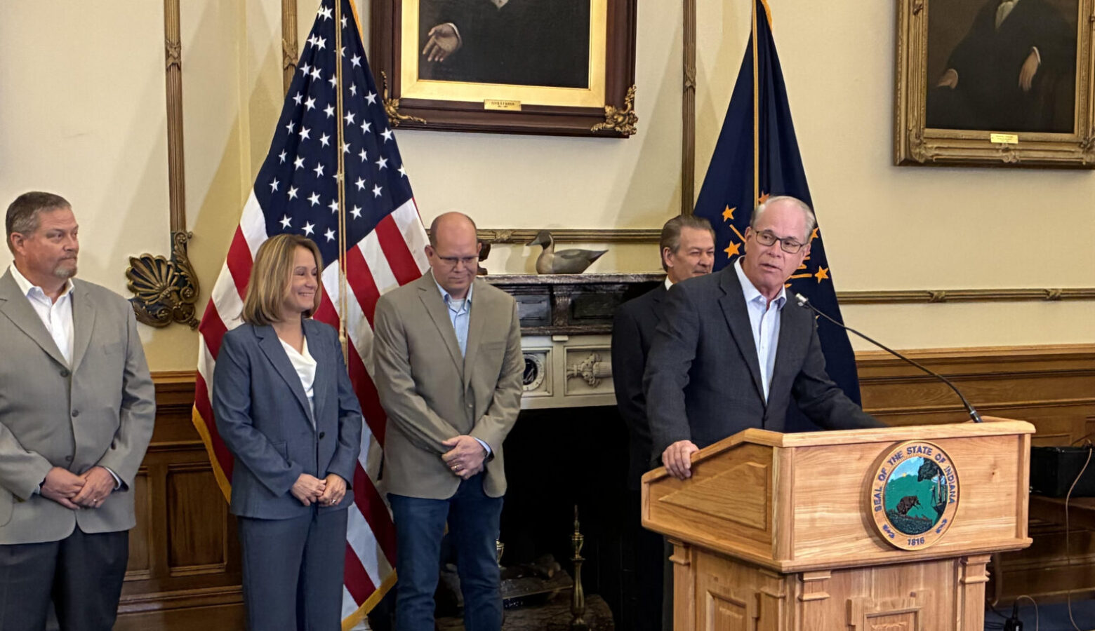 Governor Mike Braun stands at a podium, speaking into a microphone. Several people stand behind him, flanked by the American flag and Indiana flag.
