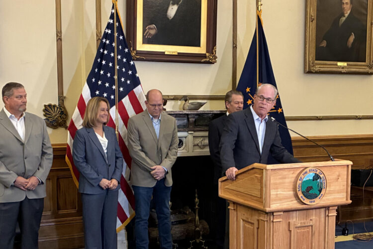 Governor Mike Braun stands at a podium, speaking into a microphone. Several people stand behind him, flanked by the American flag and Indiana flag.