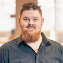 Headshot of James Edwards. He has short, curly red hair and is wearing a black button up open at the collar to reveal a gold chain.