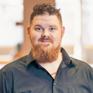 Headshot of James Edwards. He has short, curly red hair and is wearing a black button up open at the collar to reveal a gold chain.