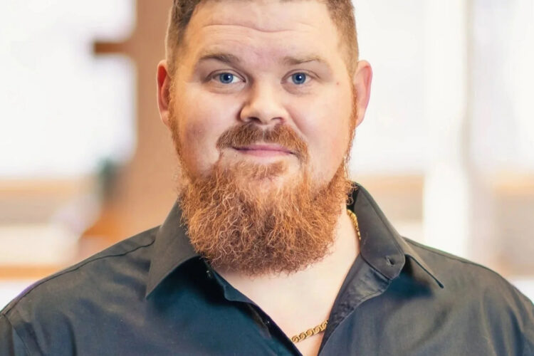 Headshot of James Edwards. He has short, curly red hair and is wearing a black button up open at the collar to reveal a gold chain.