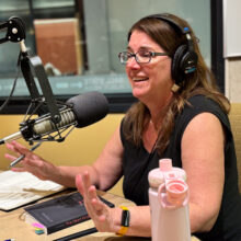 Jill Christman sits in a radio studio in front of a microphone, wearing headphones. She is smiling and talking.