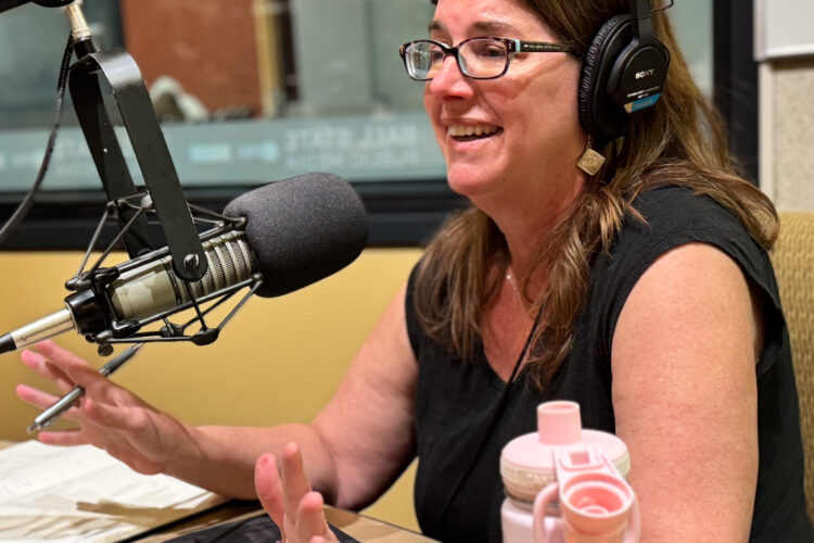 Jill Christman sits in a radio studio in front of a microphone, wearing headphones. She is smiling and talking.