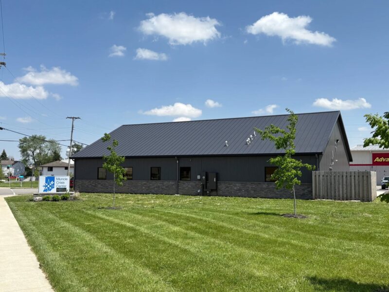A large black building sits on a green lawn. A sign next to it reads "Muncie Crisis Center - Meridian Health Services."