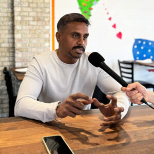 A man in a long sleeve shirt sits at a table talking; he is gesturing with his hands. A person out of frame is holding a microphone up to his mouth.