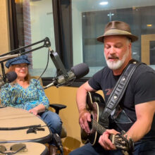 Ruthie Berkey and Randy Eyestone are sitting in a radio studio, Ruthie is watching Randy play guitar and sing.
