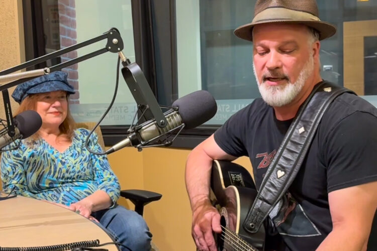 Ruthie Berkey and Randy Eyestone are sitting in a radio studio, Ruthie is watching Randy play guitar and sing.