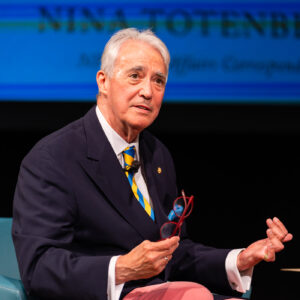 Scott Simon sits on stage in a suit, holding a pair of glasses. A large projection behind him partially reads "NINA TOTENBERG."