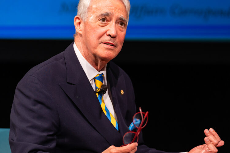 Scott Simon sits on stage in a suit, holding a pair of glasses. A large projection behind him partially reads "NINA TOTENBERG."