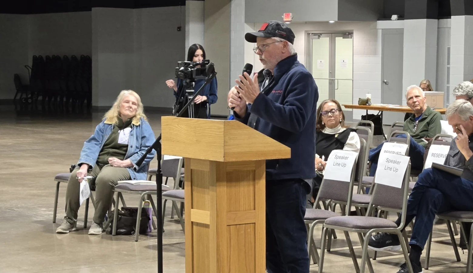 A man stands at a podium talking into a microphone at a public meeting. A small crowd of people sits behind him.