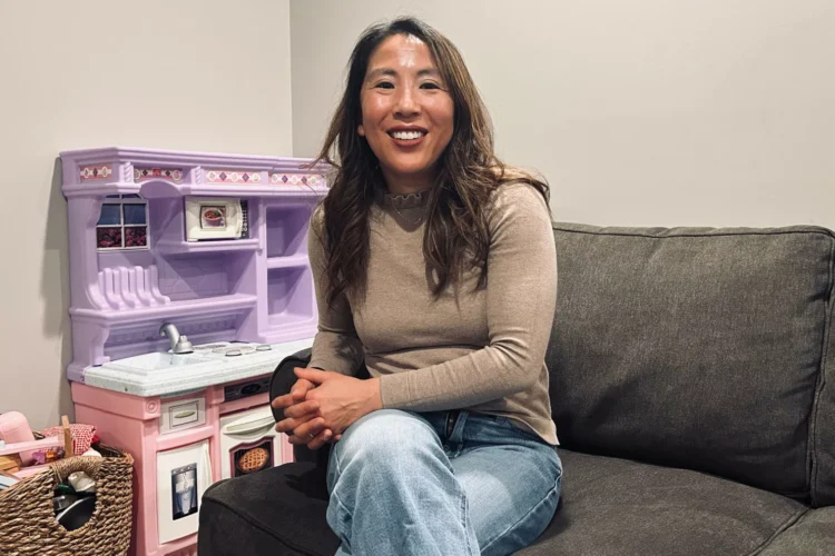 Jamie Potter sits with legs crossed on a couch next to a basket of toys and a children's toy kitchen.