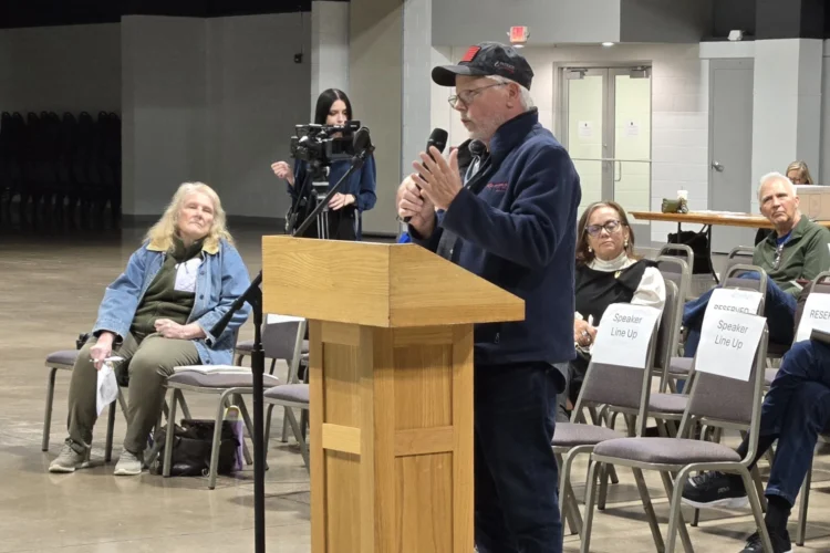 A man stands at a podium talking into a microphone at a public meeting. A small crowd of people sits behind him.