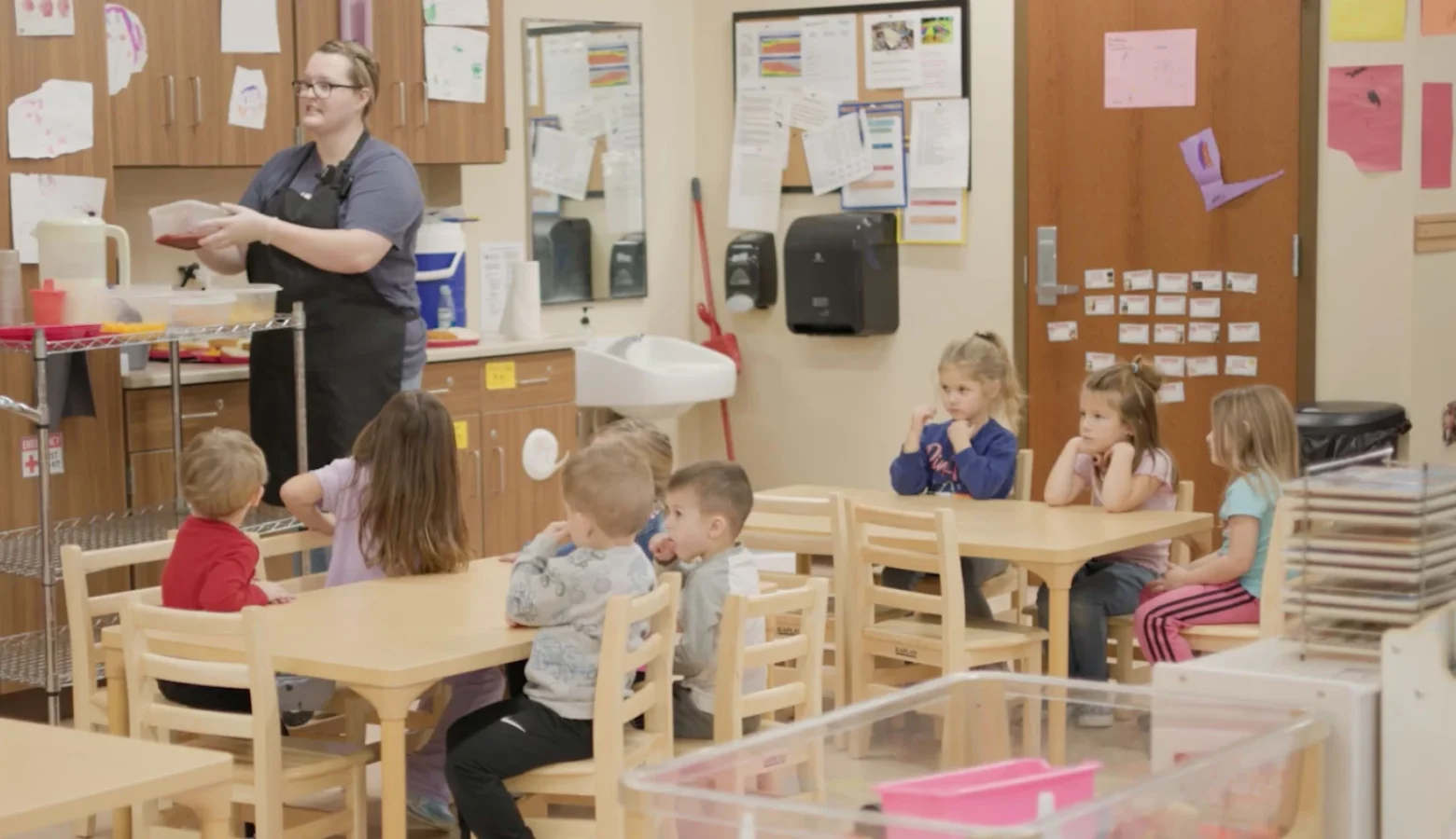 A photo of a preschool classroom with seven children seated around tables and a teacher standing nearby with what appears to be a snack cart.