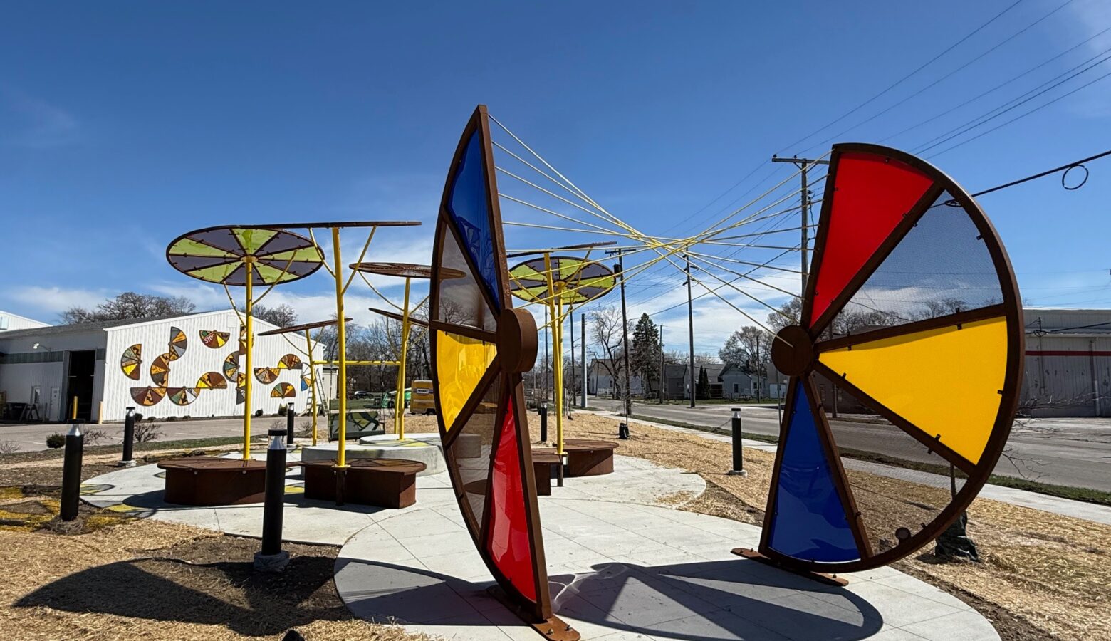 A sculpture features two large half-wheels of steel and colored glass - red, blue, and yellow. The sculpture includes a seating area.