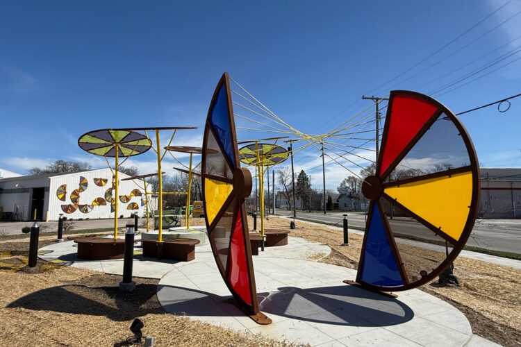 A sculpture features two large half-wheels of steel and colored glass - red, blue, and yellow. The sculpture includes a seating area.