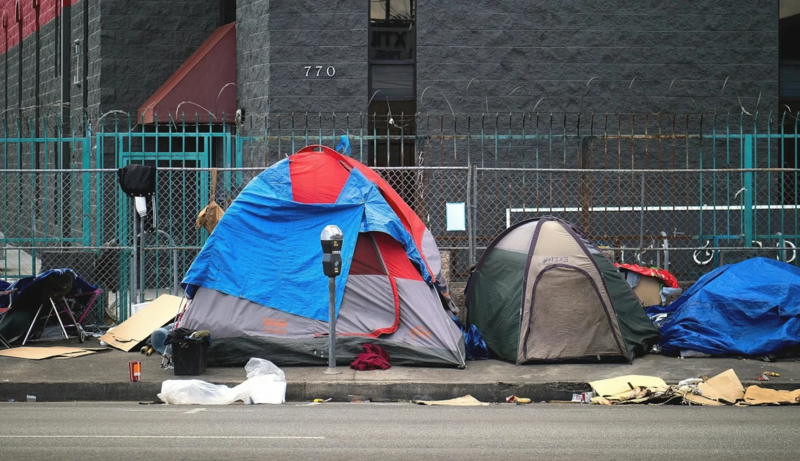 A streetside view of tents on a sidewalk near parking meters with trash strewn along the nearby curb.