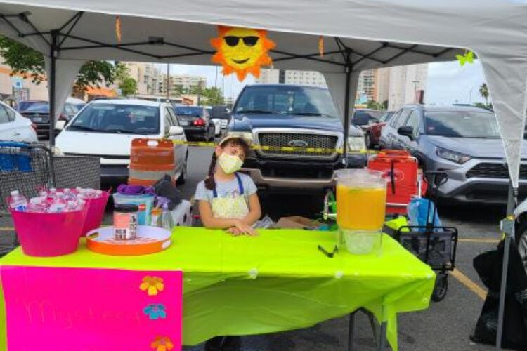 A young girl sits at a folding table underneath a conopy. A lemonade dispenser sits on the table and there is a pink sign in front of it.