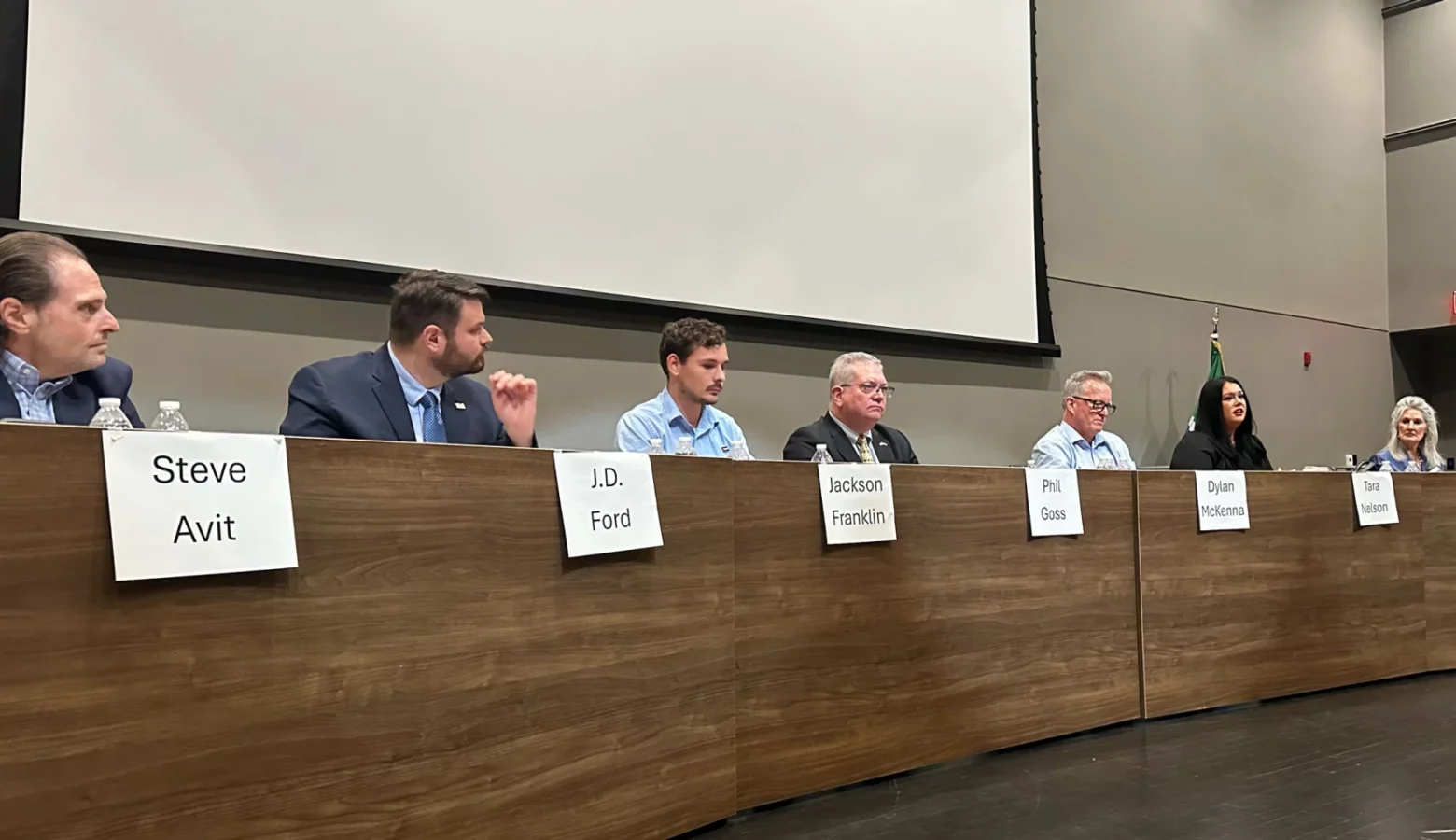 Several political candidates sit behind the same long wooden table, ready to answer questions. Each has a white nameplate.