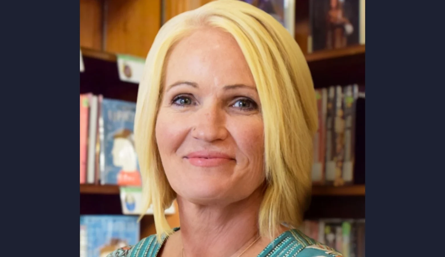 Jennifer Dorfmeyer is a white woman with short blond hair. In this headshot, she stands in front of bookshelves full of children's books.
