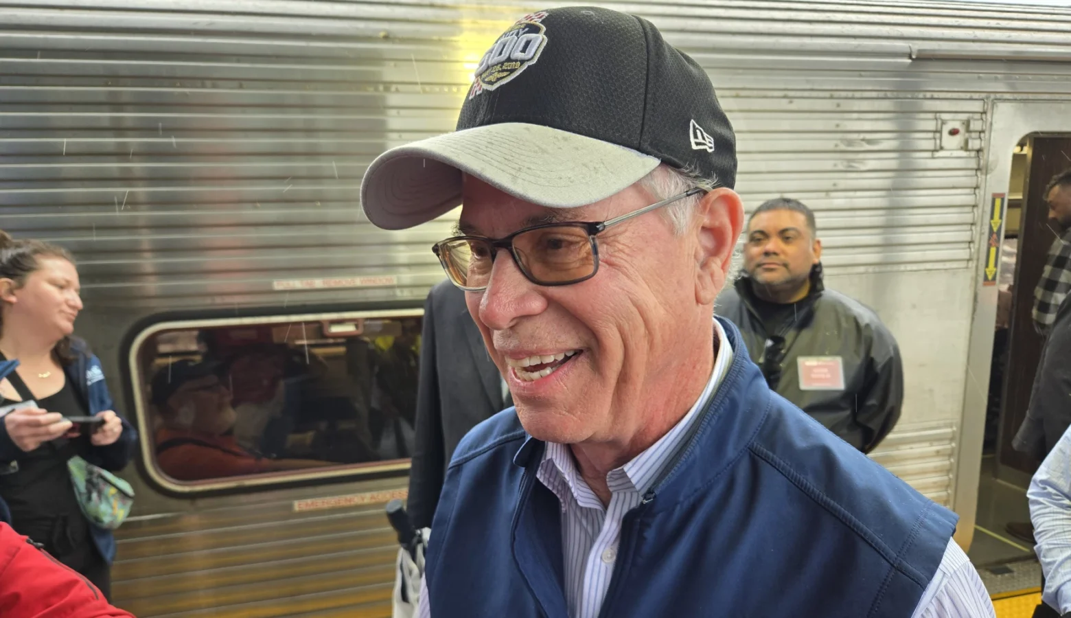 Governor Mike Braun stands in front of a train car. He is a white man, wearing a baseball cap and glasses.