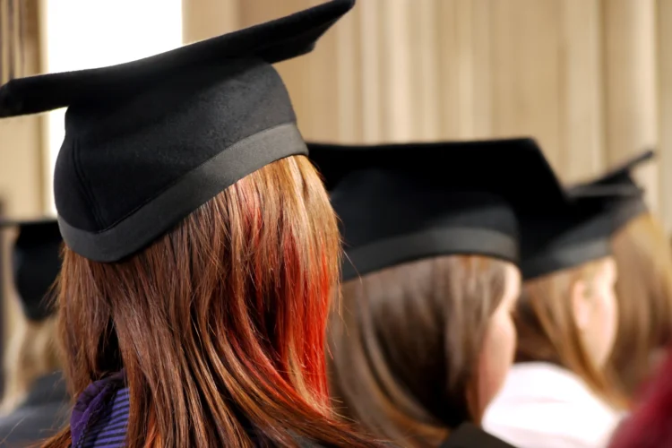 College graduates with black mortar board hats, seen from behind, sit listening to a presentation.