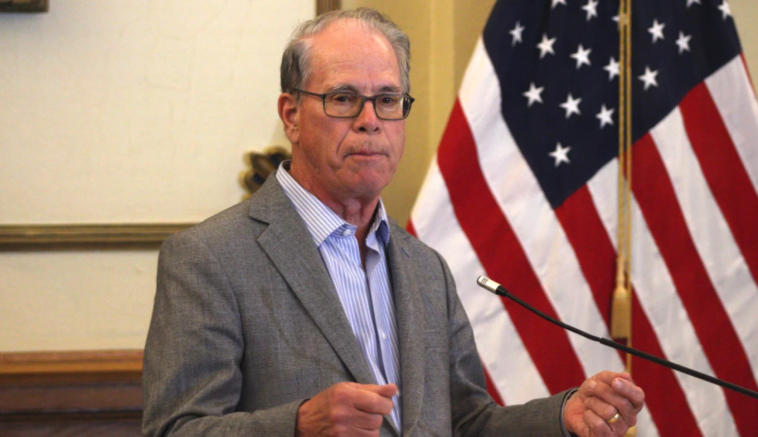 Governor Mike Braun stands behind a podium and microphone., He is a White man with glasses, wearing a gray suit jacket. He stands next to an American flag.