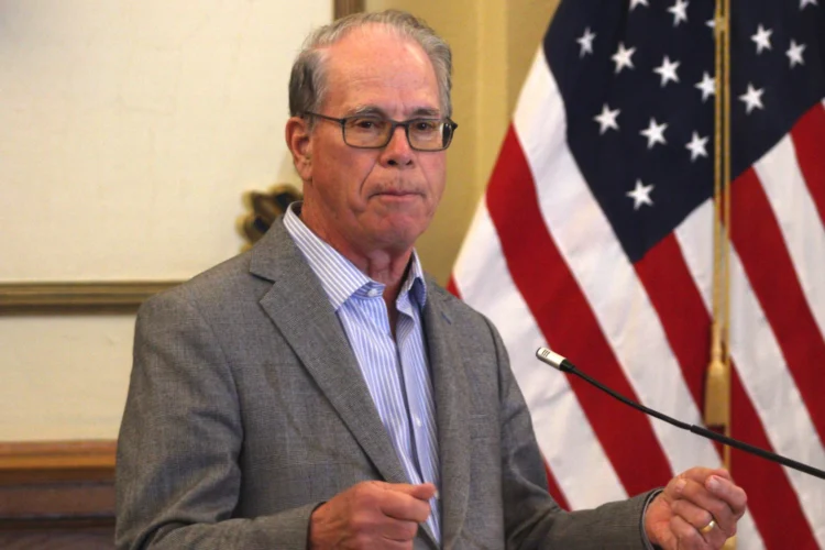 Governor Mike Braun stands behind a podium and microphone., He is a White man with glasses, wearing a gray suit jacket. He stands next to an American flag.