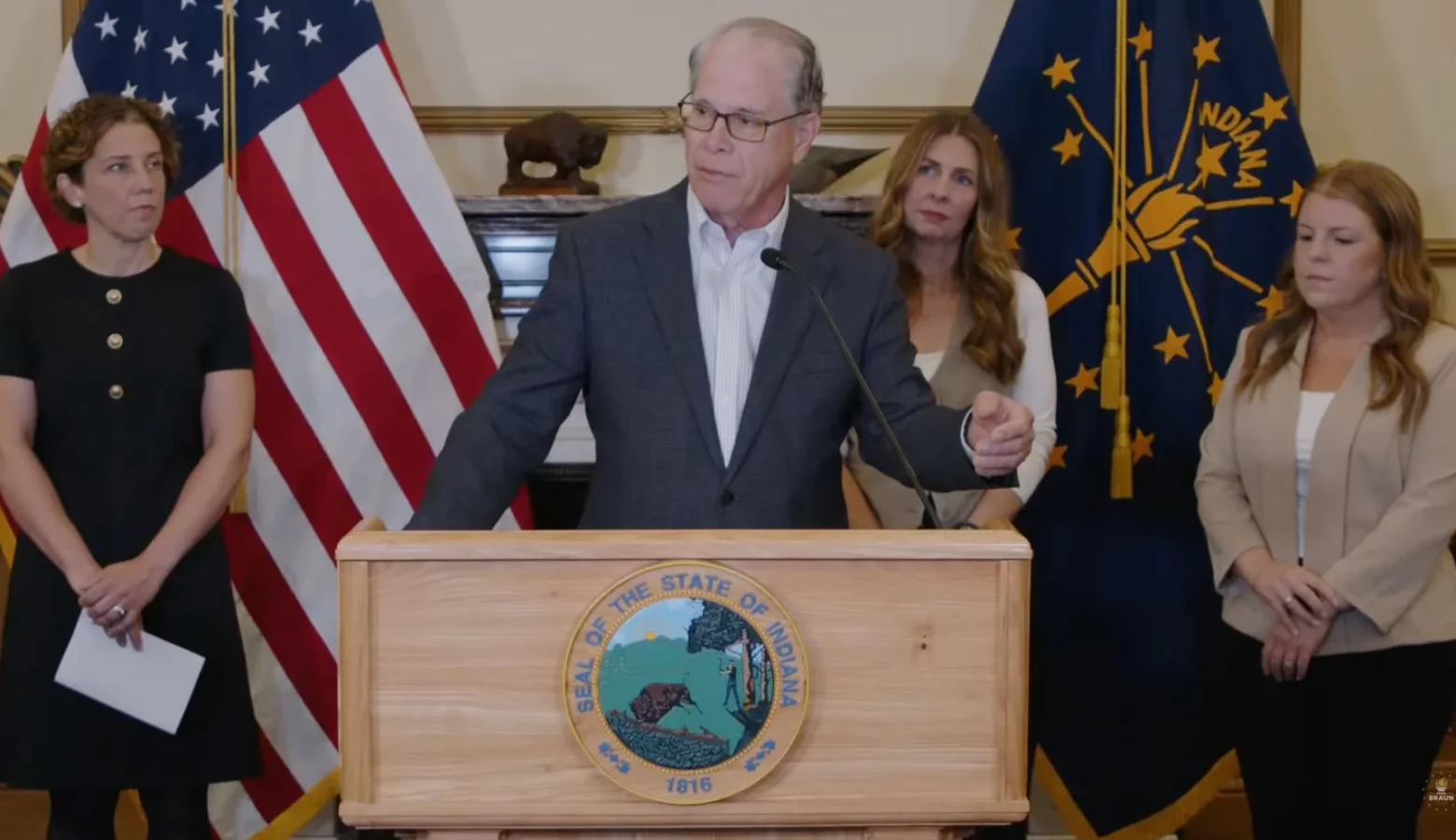 Governor Mike Braun stands behind a podium and microphone., He is a White man with glasses, wearing a gray suit jacket. He is surrounded by several women and the American flag and the Indiana flag.