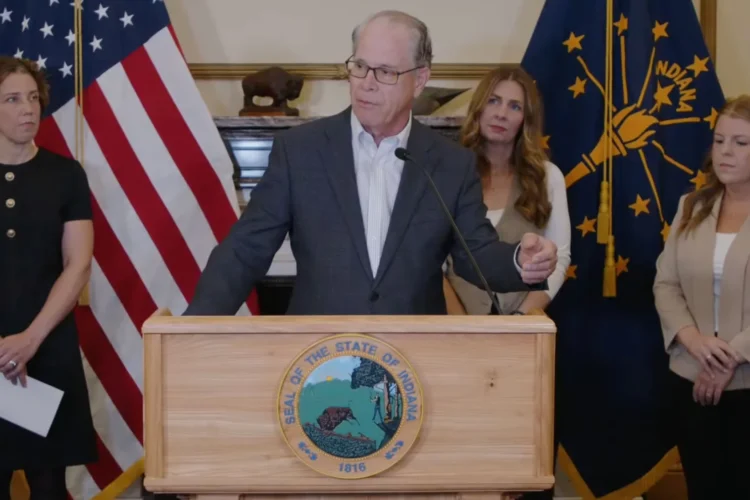 Governor Mike Braun stands behind a podium and microphone., He is a White man with glasses, wearing a gray suit jacket. He is surrounded by several women and the American flag and the Indiana flag.