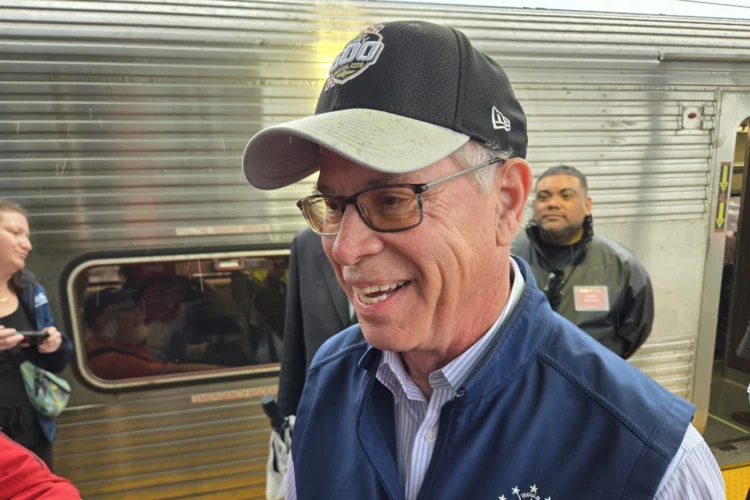 Governor Mike Braun stands in front of a train car. He is a white man, wearing a baseball cap and glasses.