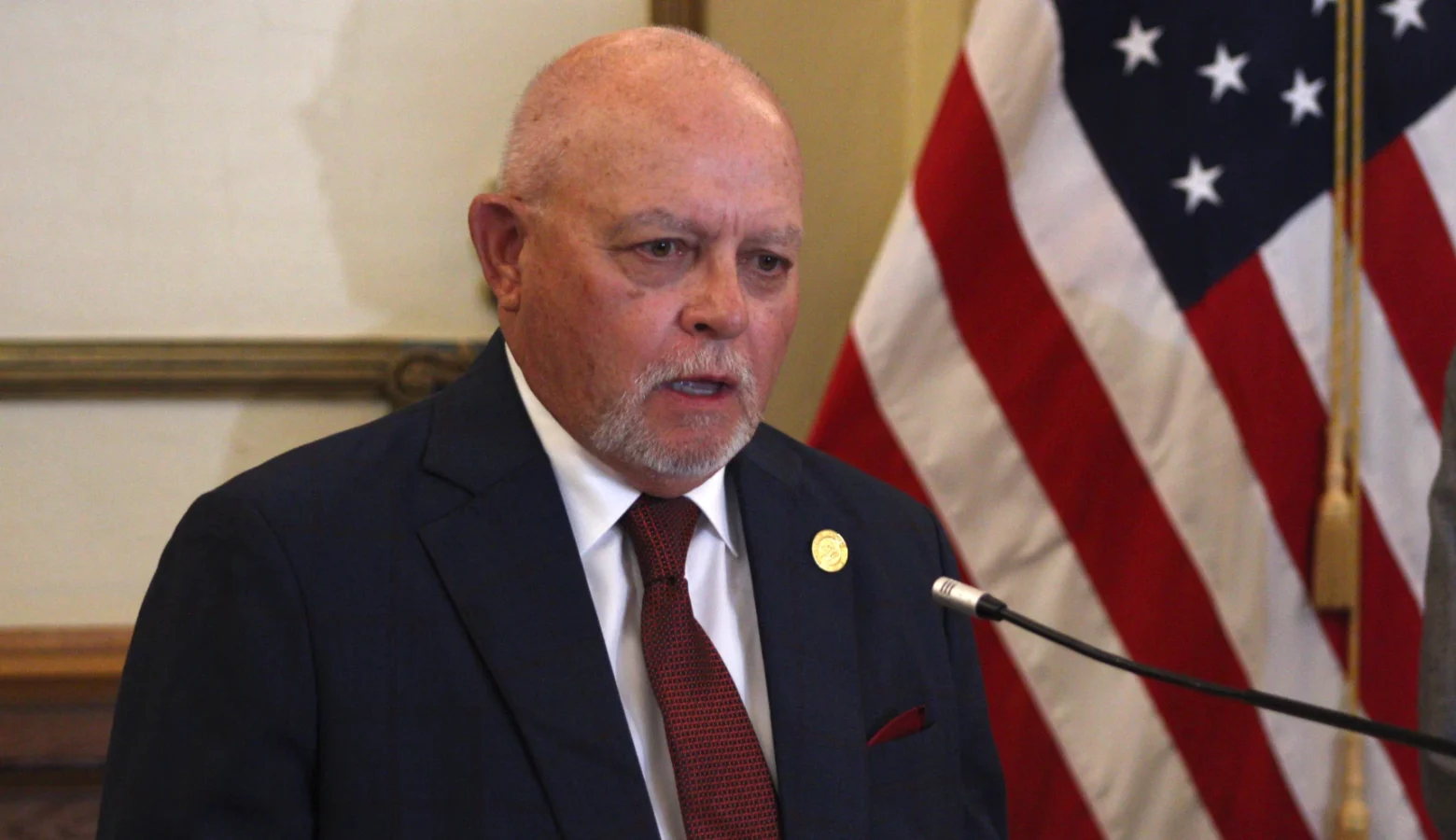 Rep. Doug Miller stands in front of a podium, speaking into a microphone. He is a bald White man wearing a dark suit and red tie.