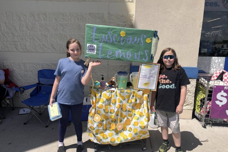 Two kids stand on each side of a small lemonade stand. A sign fixed to the top of the stand reads "Luscious Lemons." Two full pitchers rest on the stand one yellow and one a light blue.