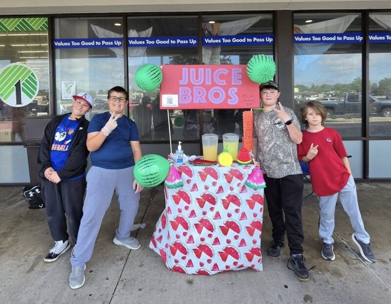 A colorful lemonade stand sits between a group of 4 kids. A sign fixed to the top of the stand says "Juice Bros." The kids pose for a picture