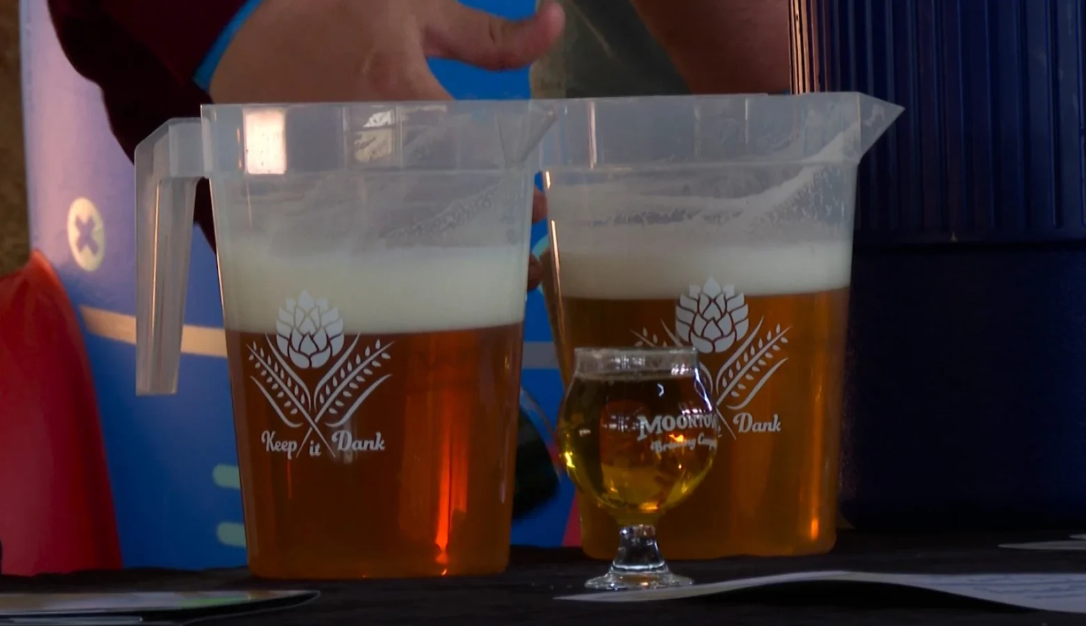 A closeup shot of two half full, plastic beer pitchers with a small goblet filled with beer in front of them. A server stands behind the containers.
