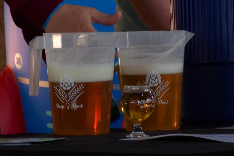 A closeup shot of two half full, plastic beer pitchers with a small goblet filled with beer in front of them. A server stands behind the containers.